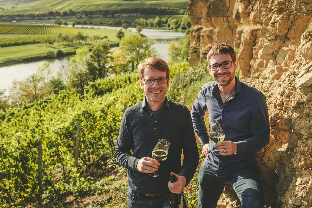 Timo und Matthias Sauerwein mit Weingläsern vor einer Muschelkalkwand in den Weinbergen von SAUERWEINS an der südlichen Mosel
